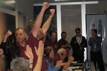 In this image, a group of men and women are celebrating the successful orbit insertion of Mars Reconnaissance Orbiter on March 10, 2006.  Mission manager Jim Graf is featured.  He is a Caucasian man in his fifties with  brown hair and a salt and pepper goatee.  His arms are raised in celebration.  Behind him is the Director of NASA's Jet Propulsion Laboratory, Dr. Charles Elachi.  He is a  man in his fifties with thinning brown and gray hair and glasses.  They are both wearing maroon-colored short-sleeved shirts with mission emblems on them.  Dr. Elachi has a brown leather jacket on also.  They are surrounded by other men and women clapping in mission control.